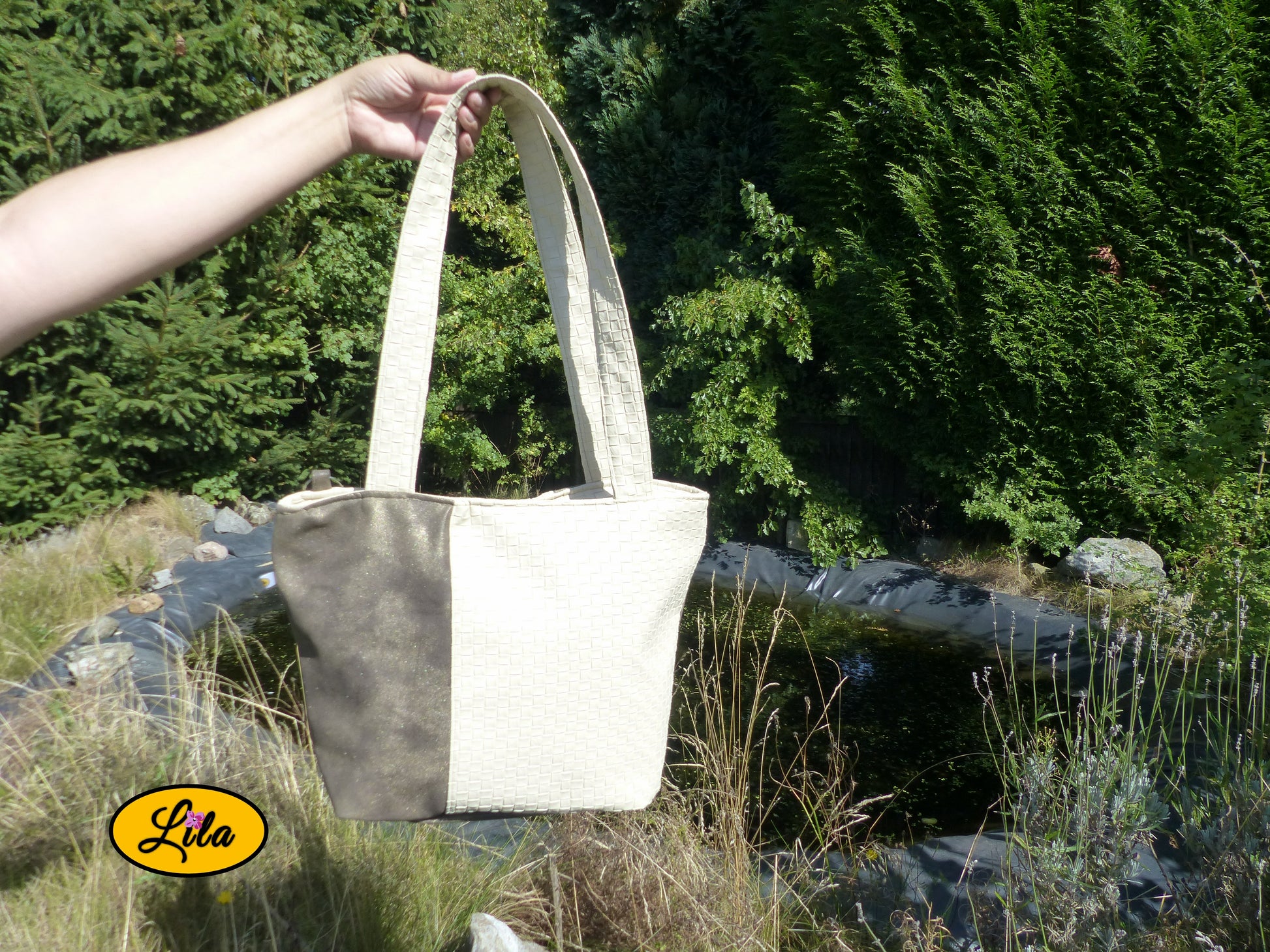 Sac 4 saisons en simili cuir blanc crème texturé et brun doré porté à la main devant un jardin et un étang style élégant et naturel