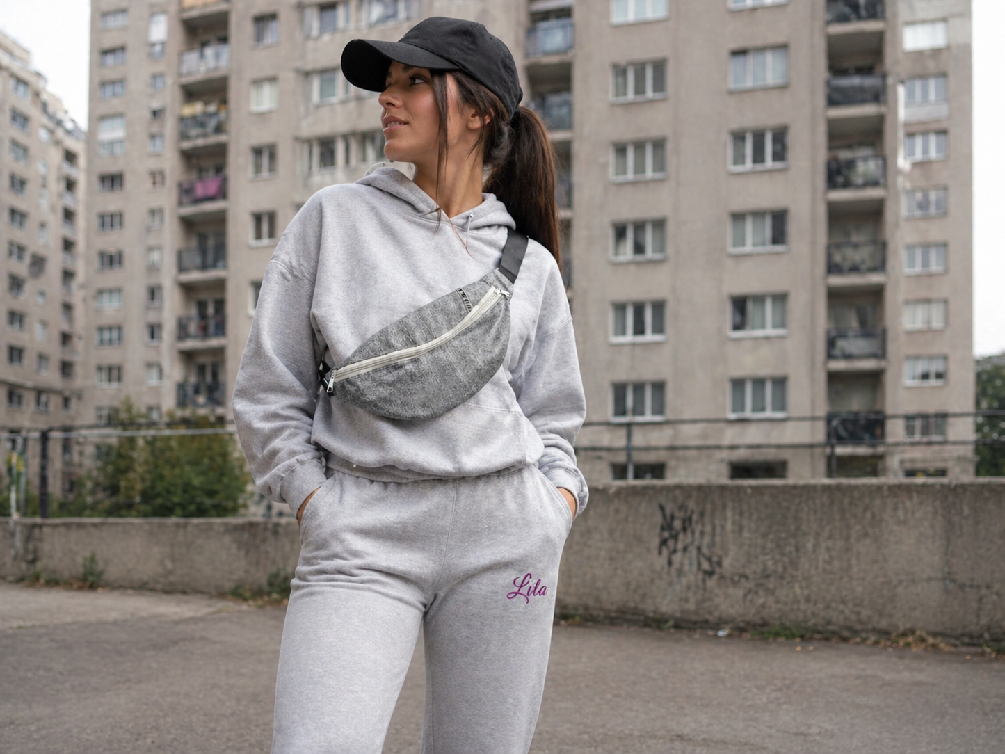 Sac banane réalisé dans une chemise noire recyclée avec sangle noire et fermeture éclair grise porté par une jeune femme en training gris et casquette noire dans une cité style urbain et streetwear