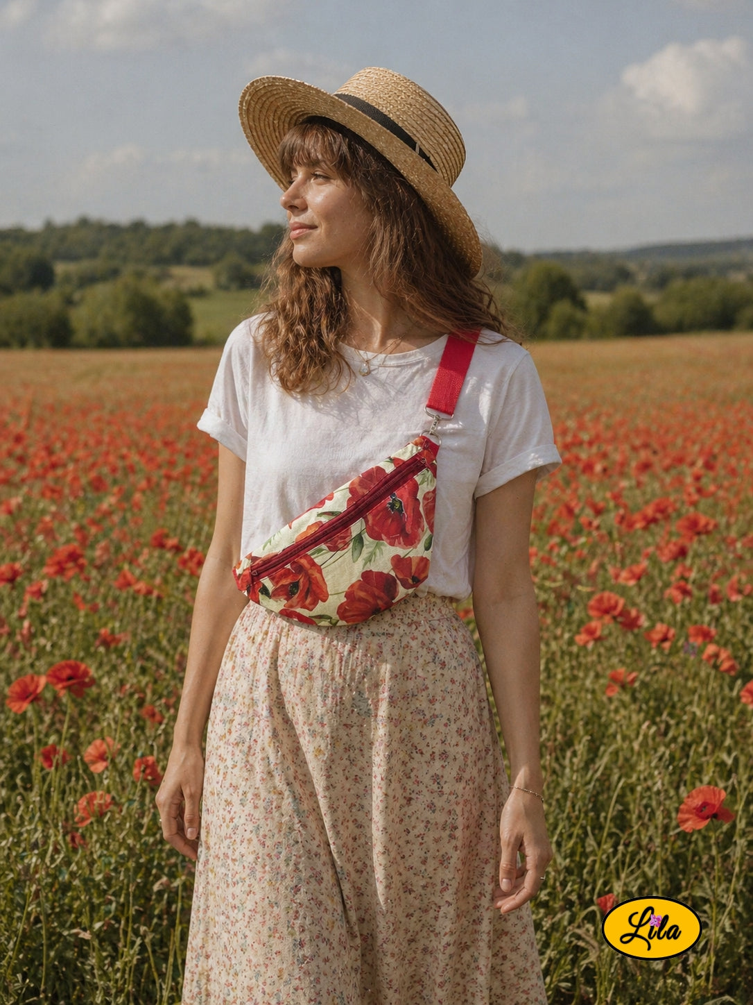 Sac banane vintage aux motifs de coquelicots avec sangle rouge et fermeture éclair bordeaux porté par une jeune femme en haut blanc et longue jupe fleurie avec chapeau de paille devant un champ de coquelicots style bohème romantique et estival