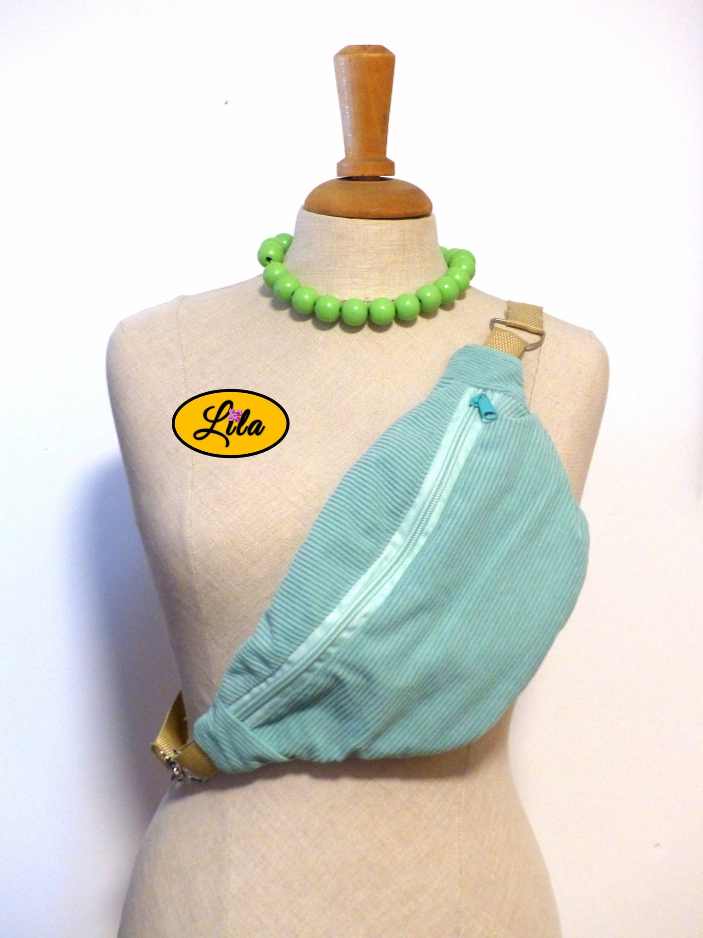Sac banane en velours côtelé vert menthe avec sangle assortie et anse couleur sable porté sur mannequin de couture sur fond blanc style tendance et artisanal