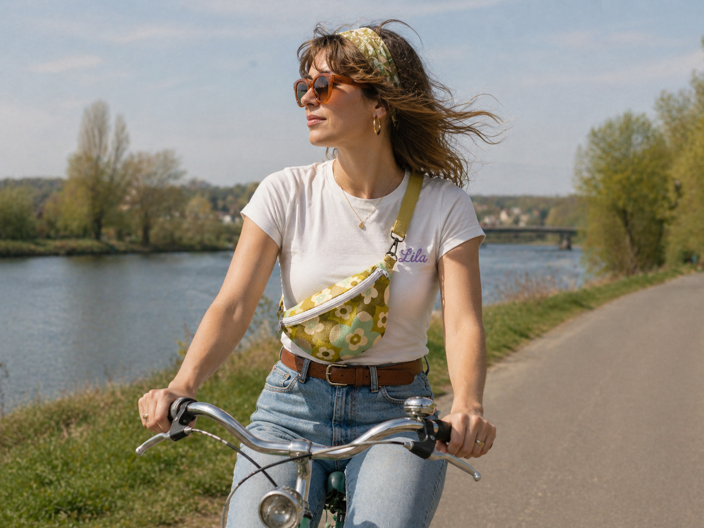 Sac banane vintage fleurie verte avec sangle kaki et fermeture éclair blanche porté par une jeune fille en jeans et t-shirt blanc roulant à vélo au bord de l’eau avec lunettes de soleil et bandeau vert dans les cheveux style bohème et estival
