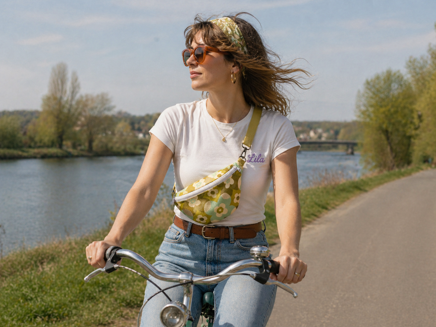 Sac banane vintage fleurie verte avec sangle kaki et fermeture éclair blanche porté par une jeune fille en jeans et t-shirt blanc roulant à vélo au bord de l’eau avec lunettes de soleil et bandeau vert dans les cheveux style bohème et estival