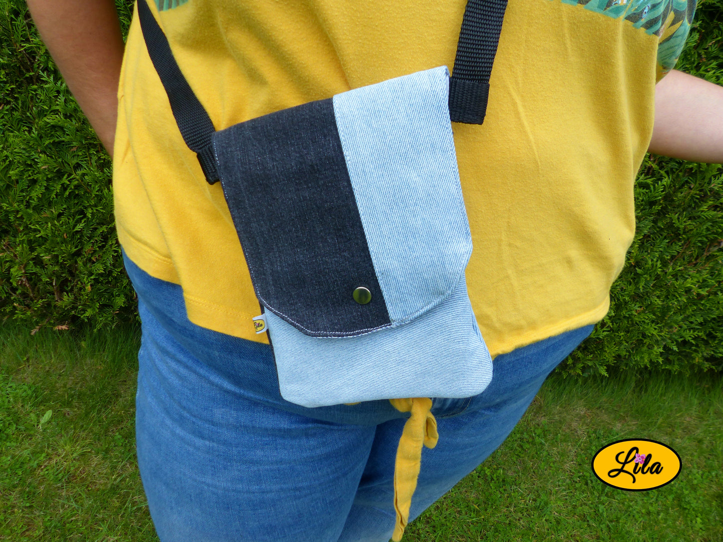 Sac bandoulière pour téléphone en jeans bleu ciel avec détails noirs porté par une jeune femme devant une haie dans un jardin avec photo cadrée sur le sac mettant en valeur son format compact et son style artisanal