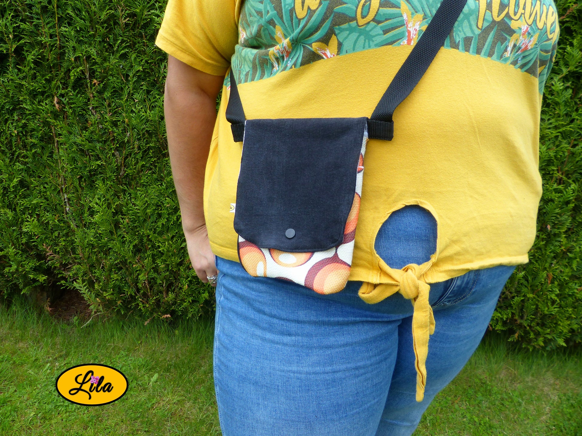 Sac bandoulière pour téléphone en jeans noir avec rabat en ancienne nappe vintage à motifs de ronds orange porté par une jeune femme devant une haie dans un jardin avec photo cadrée sur le sac mettant en valeur son style rétro et artisanal
