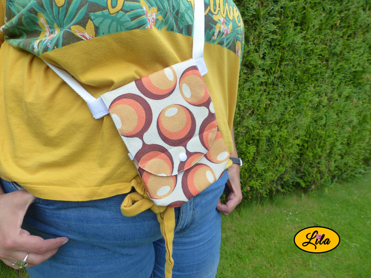 Sac bandoulière pour téléphone aux motifs vintage orange et brun, porté à la taille avec sa sangle blanche, devant une haie en extérieur