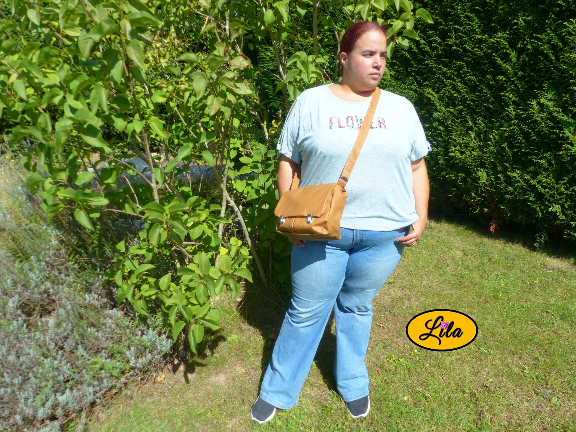 Sac besace en simili cuir brun légèrement craquelé porté en bandoulière par une femme dans un jardin près d’un arbre avec mains dans les poches du jeans vue complète style élégant et naturel