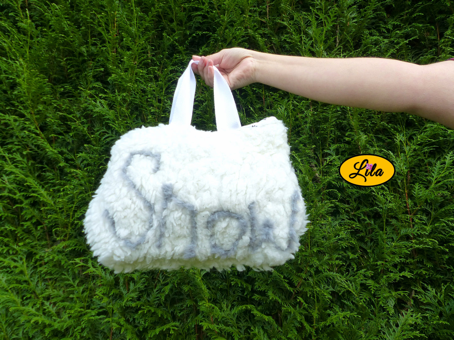 Sac à main en fausse fourrure blanche avec inscription SNOW porté à bout de bras devant une haie style doux et tendance