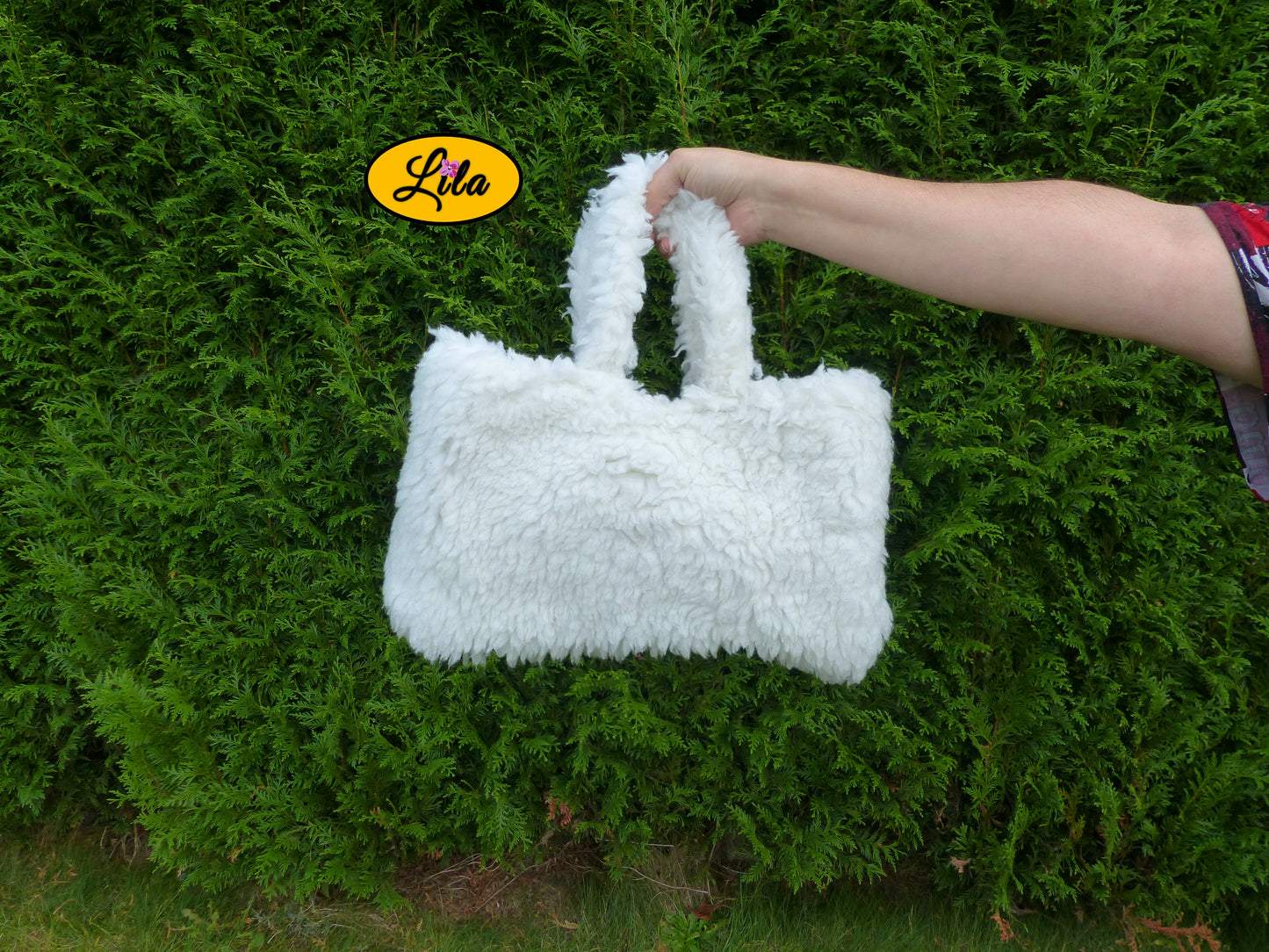 Sac à main entièrement en fausse fourrure blanche porté à bout de bras devant une haie style doux et tendance