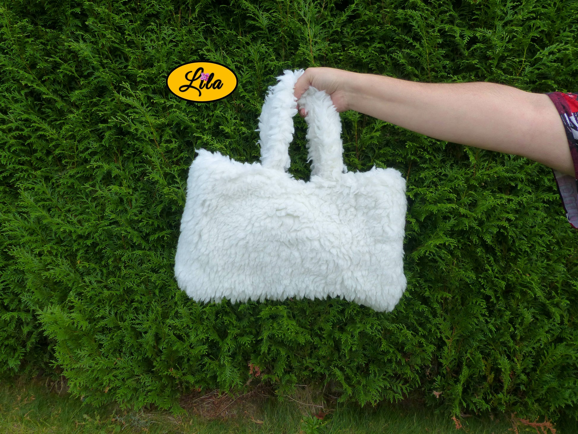 Sac à main entièrement en fausse fourrure blanche porté à bout de bras devant une haie style doux et tendance