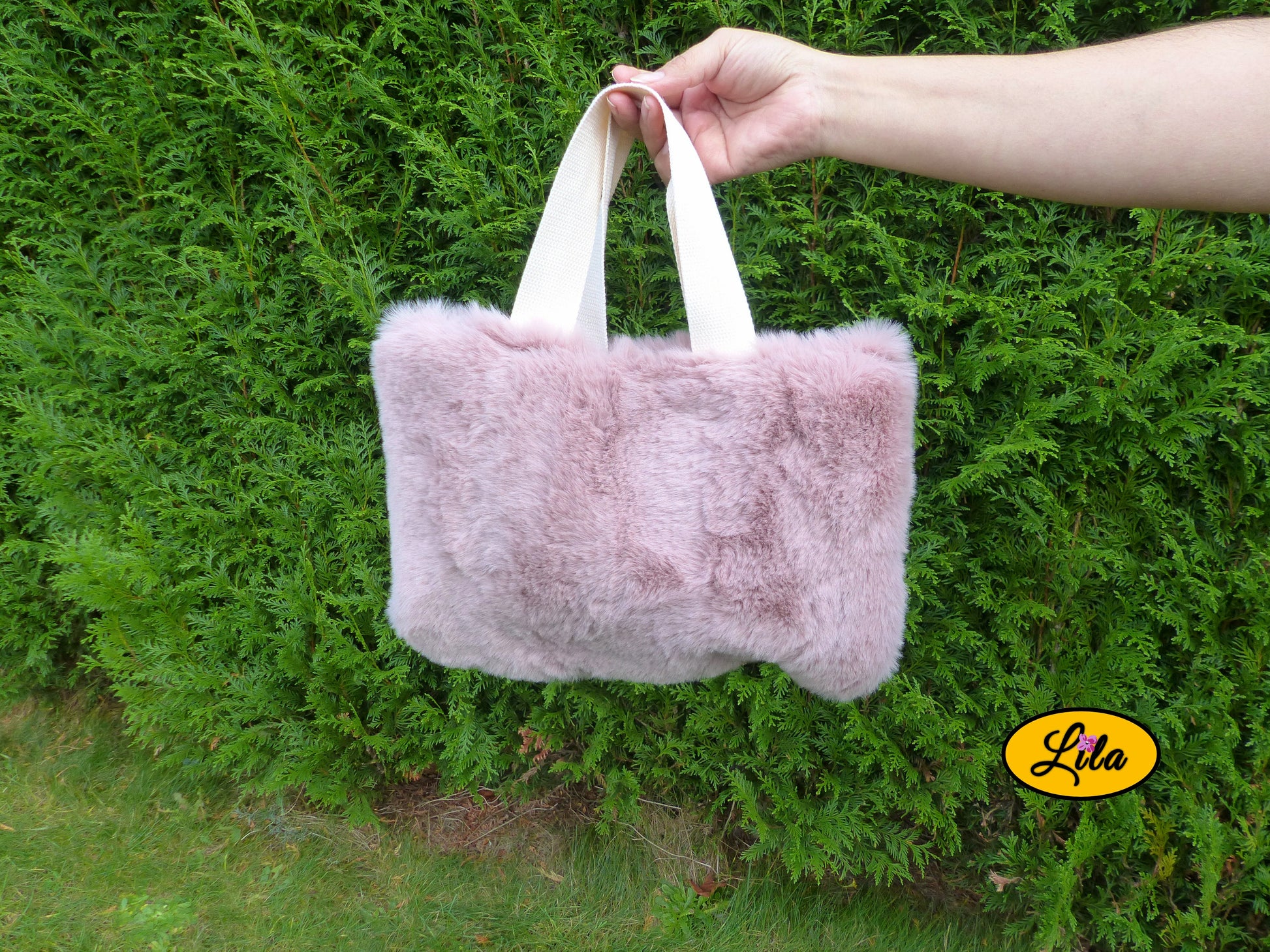 Sac à main en fausse fourrure vieux rose avec anse crème porté à bout de bras devant une haie style doux et tendance