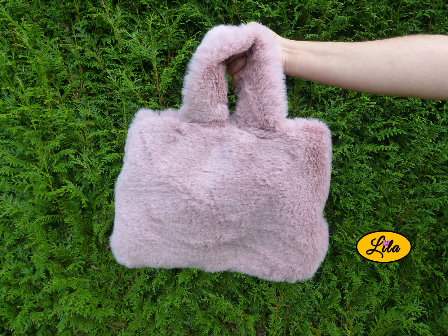 Sac à main entièrement en fausse fourrure vieux rose porté à bout de bras devant une haie style chic et cocooning