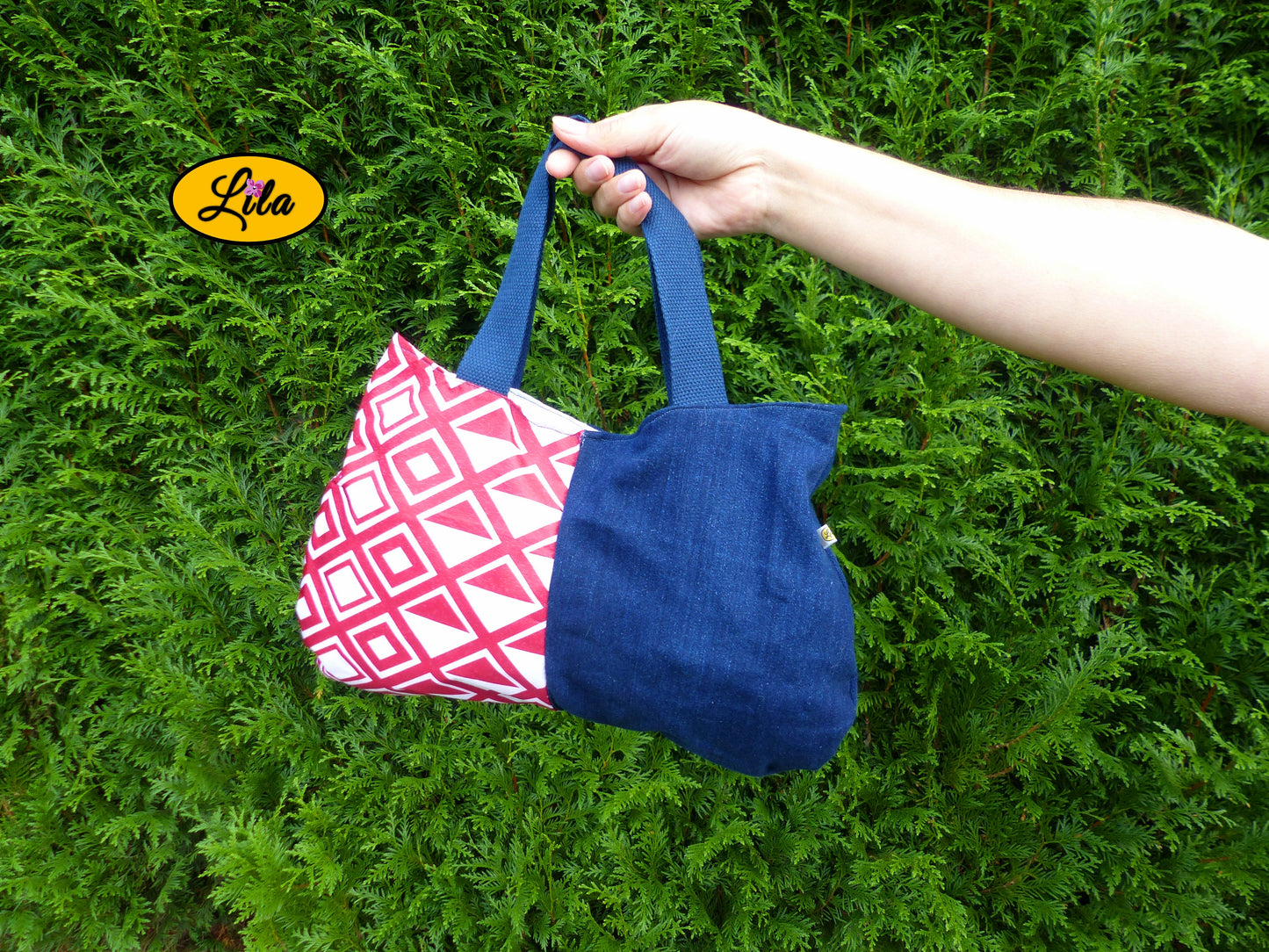 Sac à main en jeans et tissu rouge et blanc à motifs géométriques porté à bout de bras devant une haie style tendance et graphique