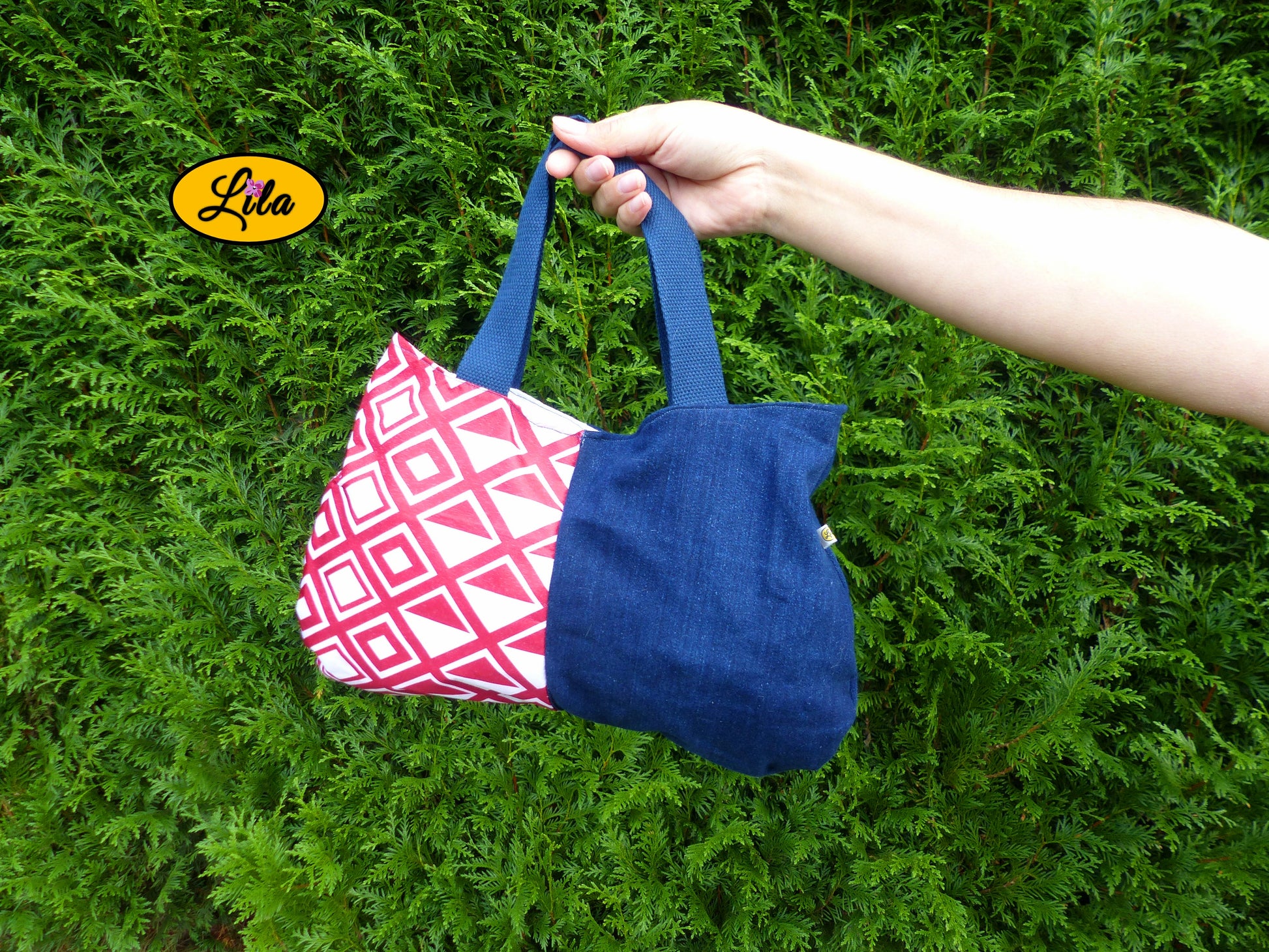 Sac à main en jeans et tissu rouge et blanc à motifs géométriques porté à bout de bras devant une haie style tendance et graphique