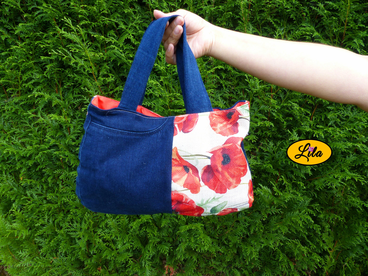 Sac à main en jeans et tissu vintage motif coquelicot porté à bout de bras devant une haie style naturel et fleuri
