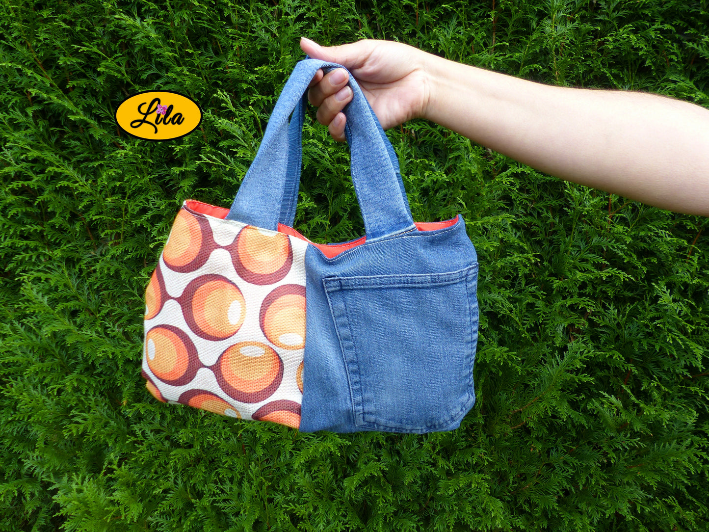 Sac à main en jeans et tissu vintage à ronds orange porté à bout de bras devant une haie style naturel et tendance