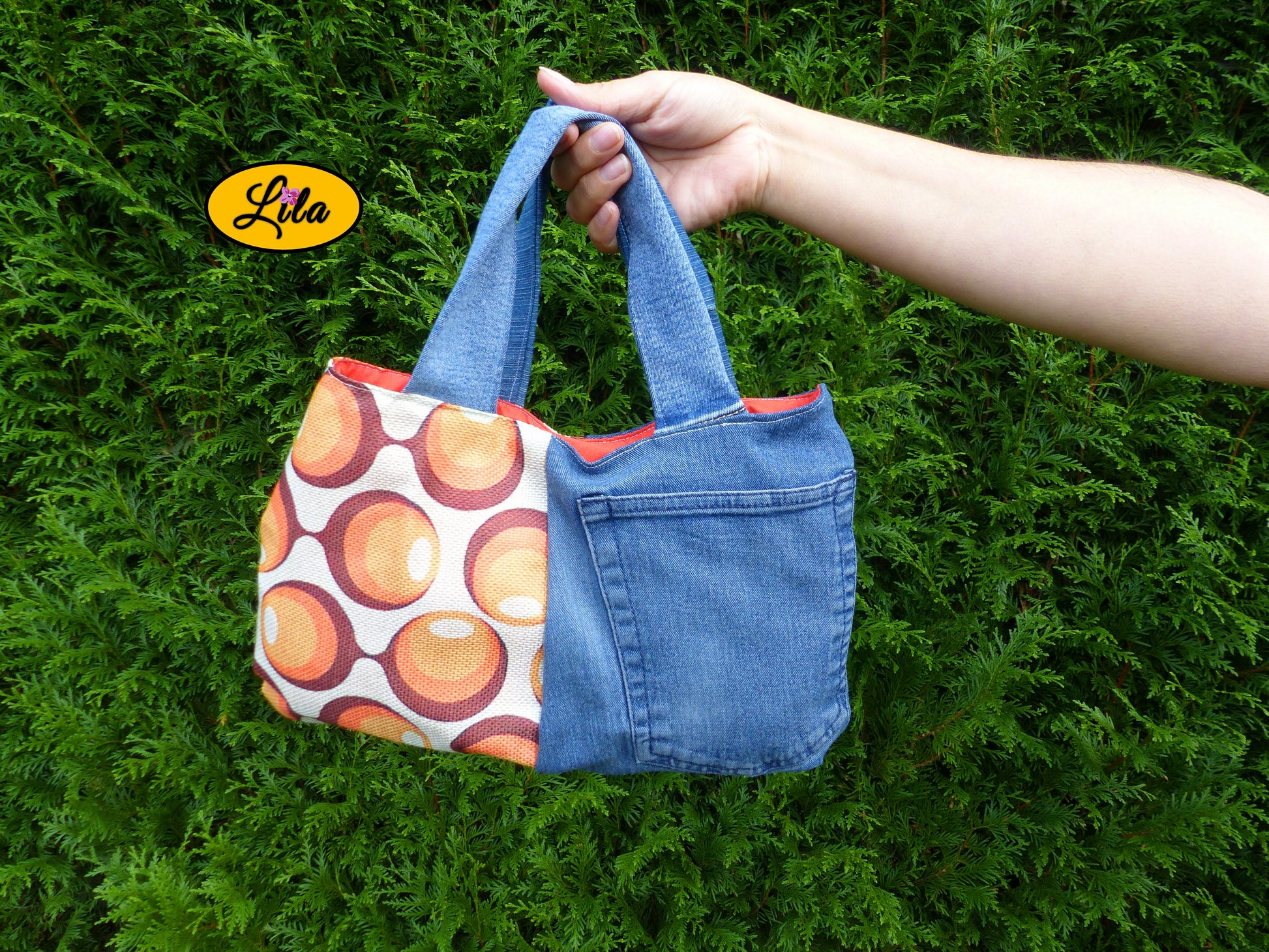 Sac à main en jeans et tissu vintage à ronds orange porté à bout de bras devant une haie style naturel et tendance