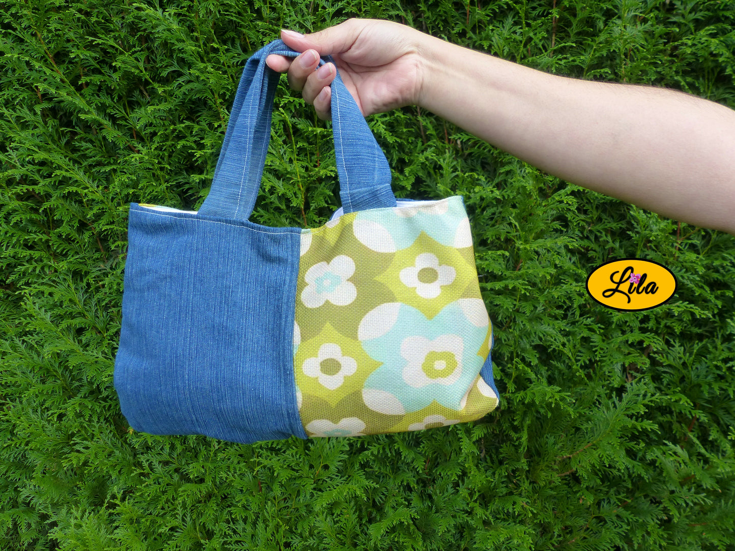 Sac à main en jeans et tissu vintage vert fleuri porté à la main devant une haie style naturel et tendance