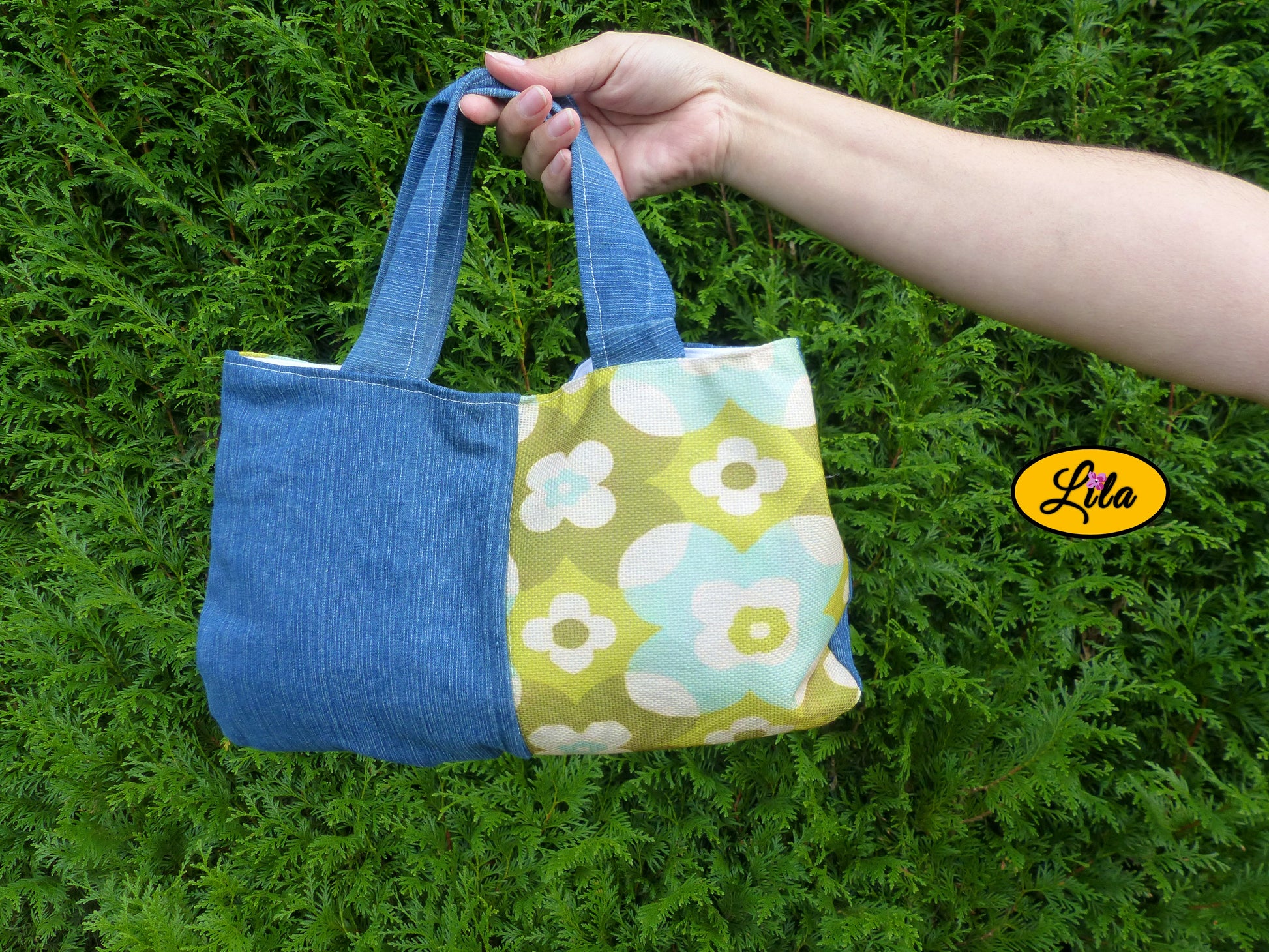 Sac à main en jeans et tissu vintage vert fleuri porté à la main devant une haie style naturel et tendance