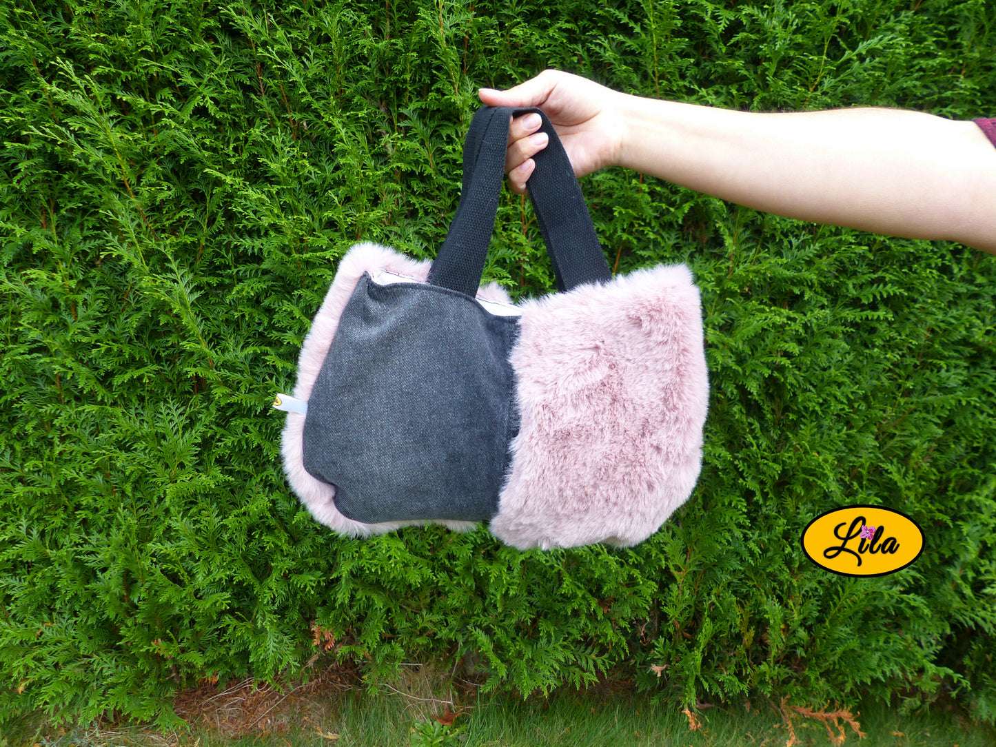 Sac à main en jeans noir et fausse fourrure vieux rose porté à bout de bras devant une haie style élégant et tendance