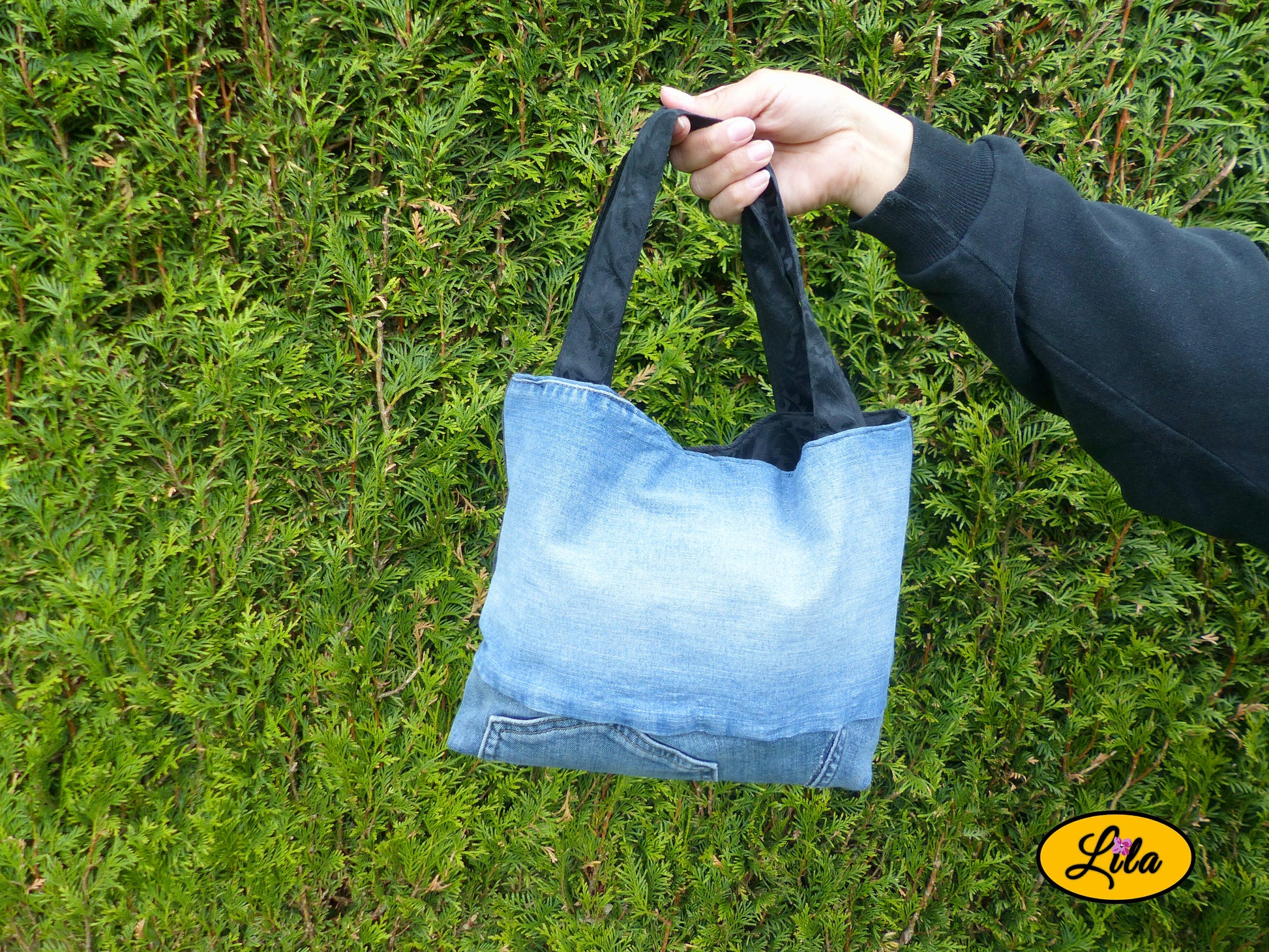 Sac patchwork en jeans recyclés bleu, bleu foncé et noir porté à bout de bras devant une haie avec anse noire à motifs floraux et fermeture à pression style upcycling et artisanal