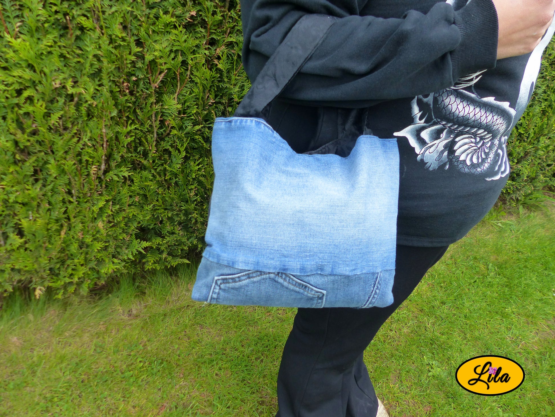 Sac artisanal réalisé à partir de trois jeans recyclés bleu, bleu foncé et noir porté par un mannequin devant une haie avec face bleue visible, anse noire à motifs floraux et fermeture à pression