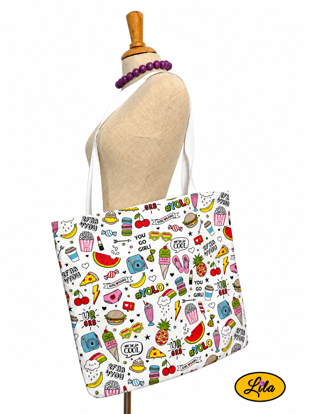 Tote bag en tissu à motifs girly avec rouges à lèvres, appareils photo et bisous porté sur mannequin de couture sur fond blanc avec sangles blanches style féminin et artisanal