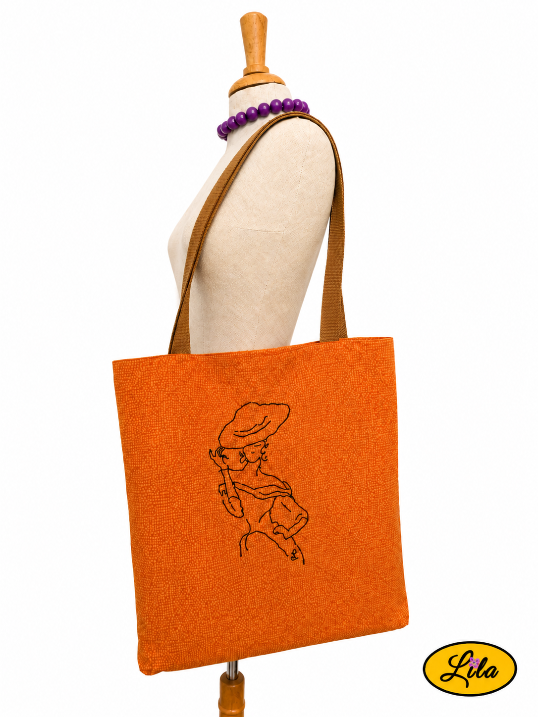 Tote bag orangé avec broderie représentant une mannequin vintage porté sur mannequin de couture sur fond blanc avec sangles couleur sable style rétro et artisanal